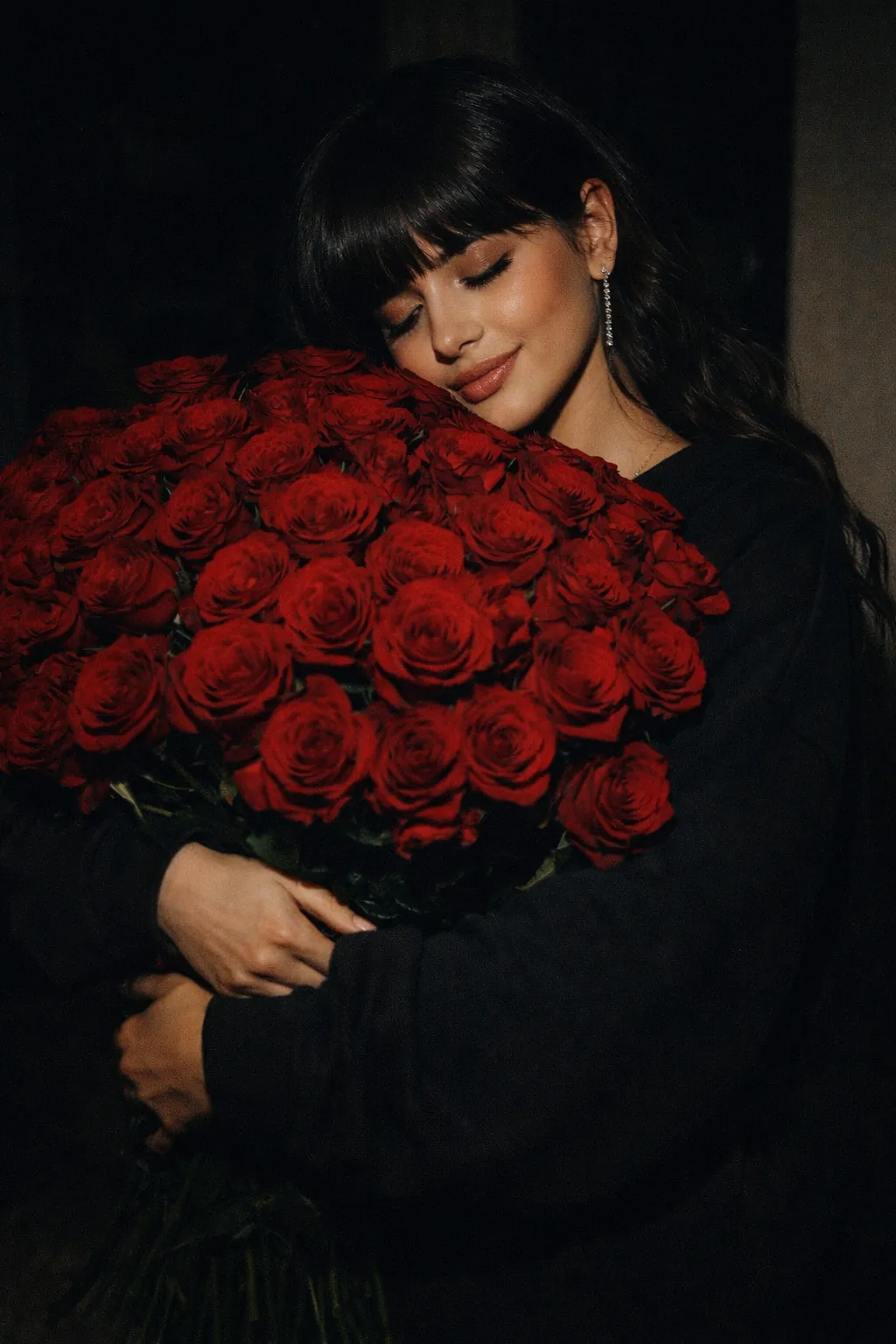Woman in oversized black sweatshirt hugging a large bouquet of red roses in moody cinematic lighting, vertical portrait.